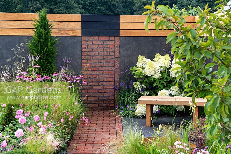 Brick path leading to a brick wall feature, wooden bench on slate tiles next to border with Hydrangea paniculata 'Phantom', Verbena bonariensis, Rosa 'Skylark' and Echinacea purpurea on left -  A Place to Ponder, RHS Tatton Park Flower Show 2018
