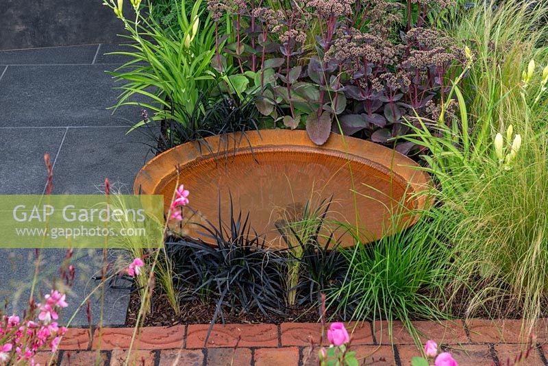 Shallow rust-effect pool in a border with Sedum 'Matrona', Ophiopogon planiscapus 'Nigrescens' and Stipa tennuissima - A Place to Ponder, RHS Tatton Park Flower Show 2018