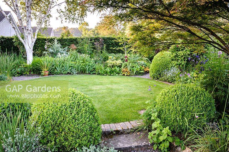 View of mixed borders surrounding circular lawn in the back garden.