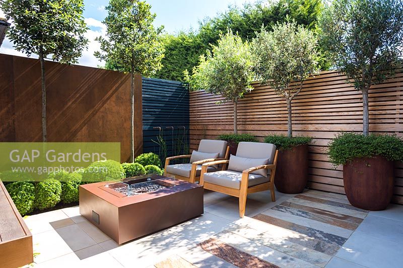 Contemporary seating area with fireplace surrounded by Olea europaea and
 Muehlenbeckia complexa in rusted steel container, Quercus ilex, 
Buxus sempervirens balls topiary by rusted panel wall and Equisetum hyemaleby 
water feature.