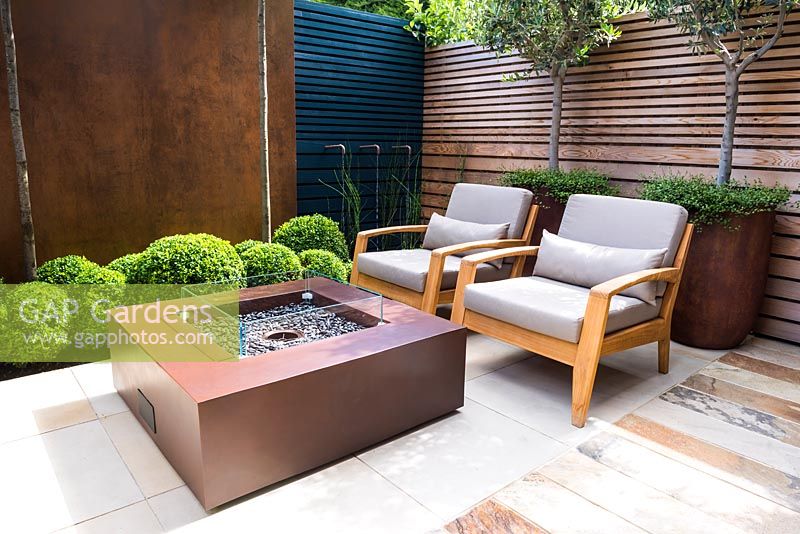 Contemporary seating area with fireplace, chairs next to big rusted corten 
steel containers with Olea europaea and Muehlenbeckia complexa and Buxus 
sempervirens balls topiary by wooden and rusted panel walls. 