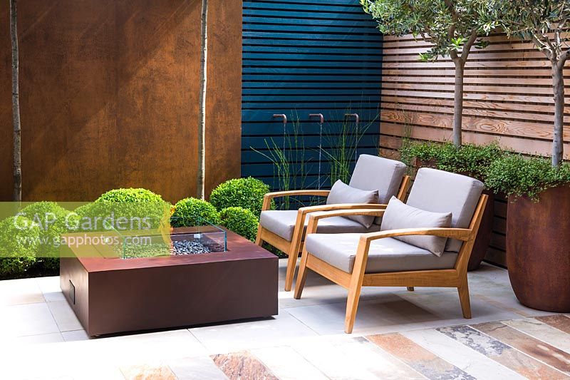 Contemporary seating area with fireplace surrounded by Olea europaea and 
Muehlenbeckia complexa in rusted steel containers, Quercus ilex, Buxus 
sempervirens balls topiary by rusted panel wall and and Equisetum hyemaleby 
water feature.