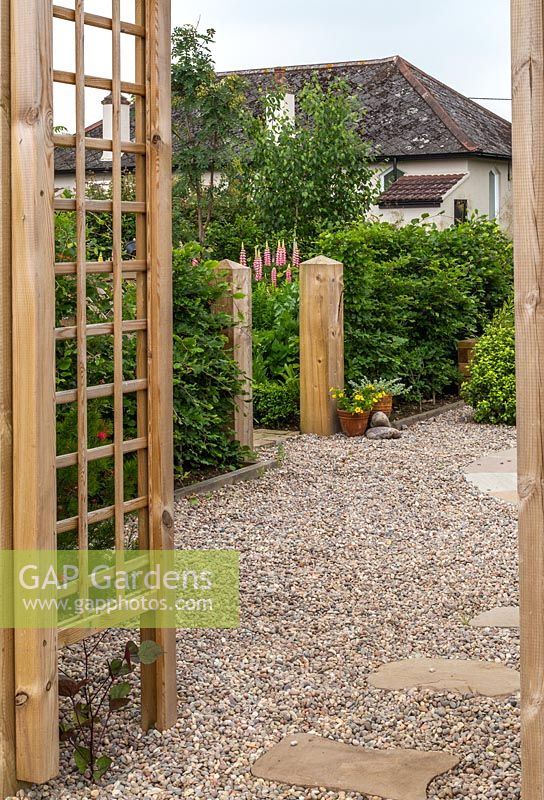 View through wooden trellis arch to gravel garden with stepping stones to garden and house beyond