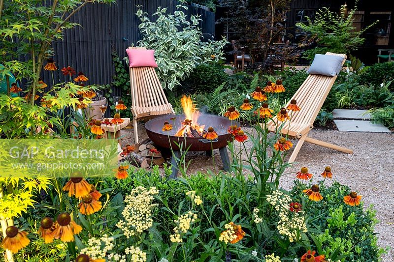 Garden designed by Nick Gough Gravel patio area with wooden chairs with logs burning in log burnerBorder planting includes:Acer palmatum Katsurabuxus sempervirens hedge Orange Helenium Moerheim Beauty Yellow Achillea Teracotta