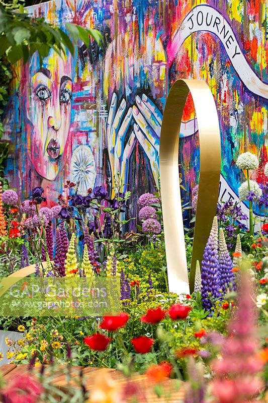 Metal ribbon sculpture against colourful grafitti wall and mixed planting. The Supershoes, Laced with Hope Garden, a partnership with Frosts. Sponsor: Frosts Garden Centres, RHS Chelsea Flower Show, 2018.