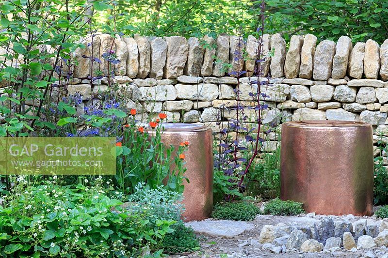 Copper seats and a fire-pit, backed by a drystone wall. The Warner Edwards Garden, a representation of Falls Farm in the Northamptonshire countryside, Sponser: Warner Edwards, RHS Chelsea Flower Show, 2018.


