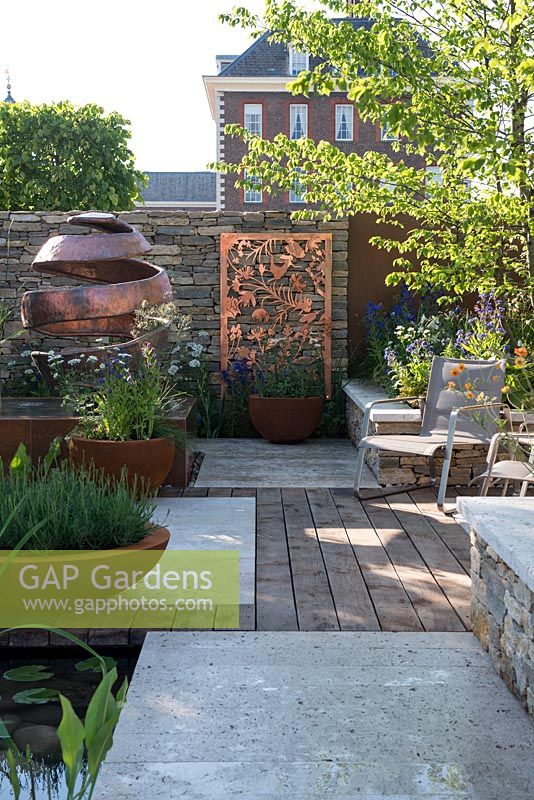 Weathered oak decking leading to Purbeck walling with copper panels, lasered to reflect the Silent Pool Gin pattern - The Silent Pool Gin Garden - Sponsor: Silent Pool Distillers - RHS Chelsea Flower Show 2018