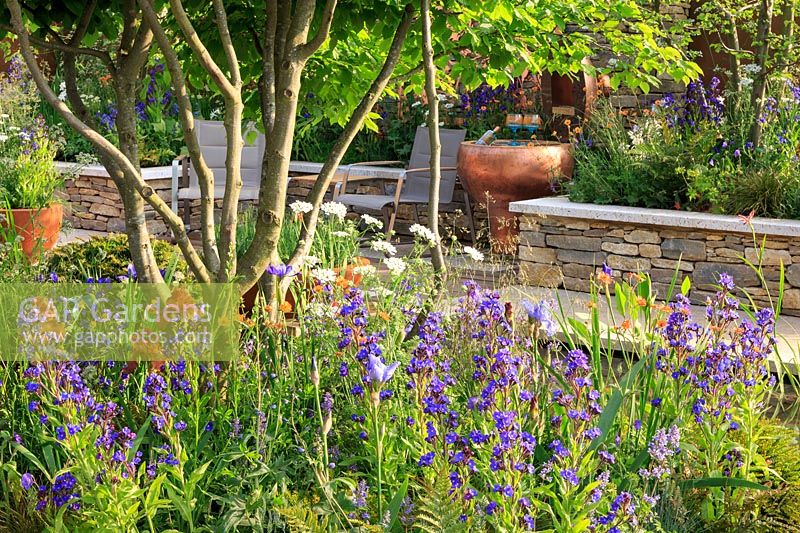 The Silent Pool Gin Garden - Seating area with copper table incorporating bottle cooler - Drystone retaining walls include Anchusa azurea 'Dropmore', Iris 'Carnival Time' and Geum 'Totally Tangerine' - Sponsor: Silent Pool Distillers - RHS Chelsea Flower Show 2018