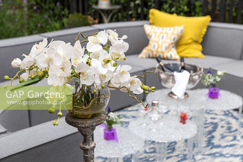 Orchid floral arrangement, with view to grey outdoor sofa arrangement and glass tables. 