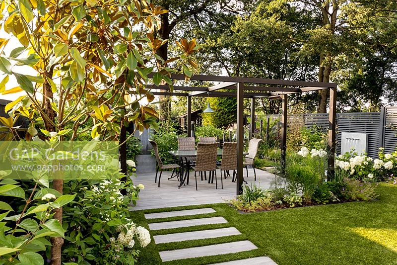 Contemporary London garden, looking towards pergola covered patio with table 
and chairs, and grey zinc water fountain and grey painted fence in background
 White flower- Hydrangea 'Annabelle' Tree on left nearest to camera Magnolia 
grandiflora Artificial lawn with stone paver path