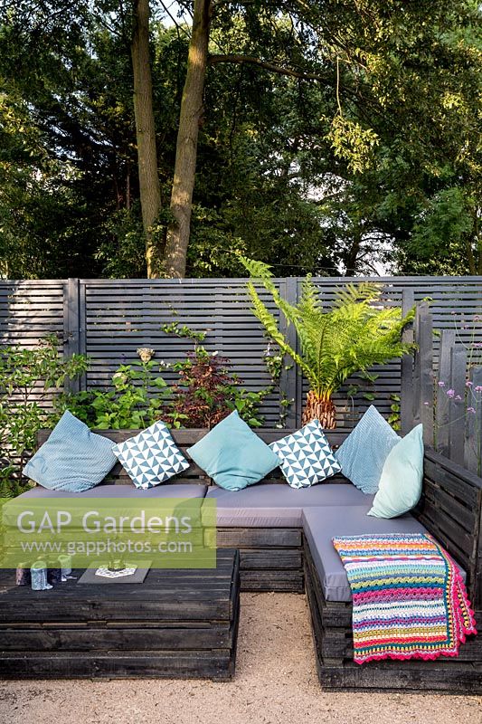 Contemporary London garden with grey painted fence. Wooden post screen - Lower seating area with decorative cushions and seat covers Purple flower -Verbena bonariensis Tree Fern Dicksonia Antarctica