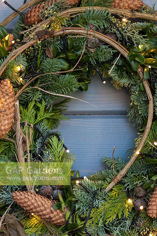 A wreath hanging on the blue wall featuring fairy lights, 
mixed greenery, and pine cones