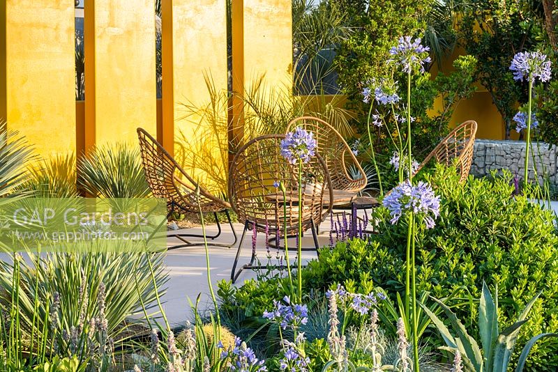 Chairs on stone terrace with yellow wall. Yucca rostrata, Agapanthus and Stachys. 'Living La Vida 120'. RHS Hampton Flower Show 2018 