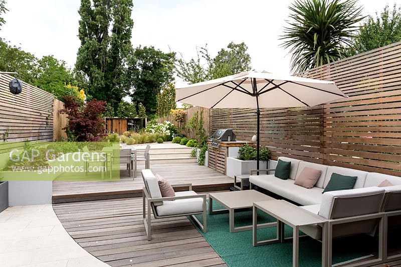 View of curved wooden decking and stone patio with garden furniture and parasol in contemporary garden. 