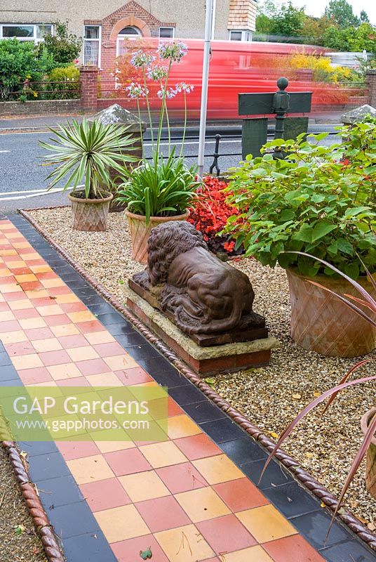 Front garden of Victorian terraced house, featuring a tiled path beside gravelled area containing statuary and planting including Begonia 'Big Boy', Agapanthus and Cordylines. The Secret Garden at Serles House, Dorset, UK. 