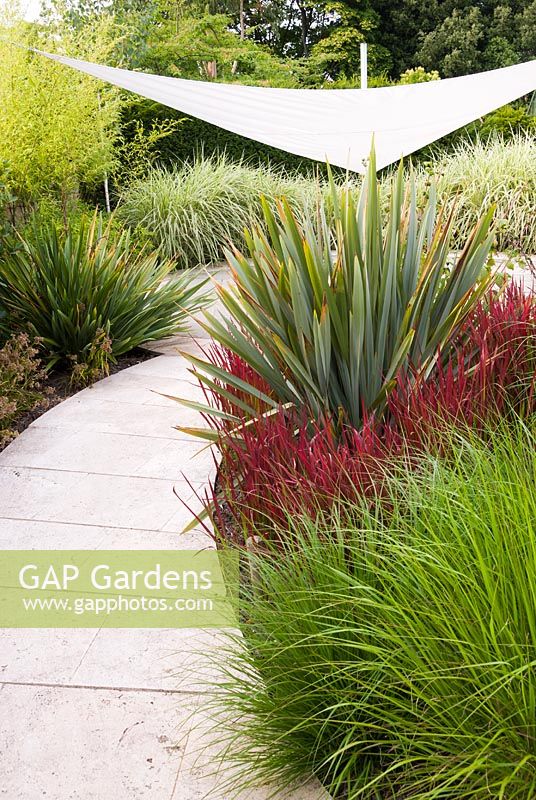 Curved paved path leads to a space shaded by white sail or awning. Nearby beds are filled with foliage interest from Phormium and ornamental grasses such as

Imperata cylindrica 'Rubra' and Pennisetum alopecuroides 'Cassian's Choice'
