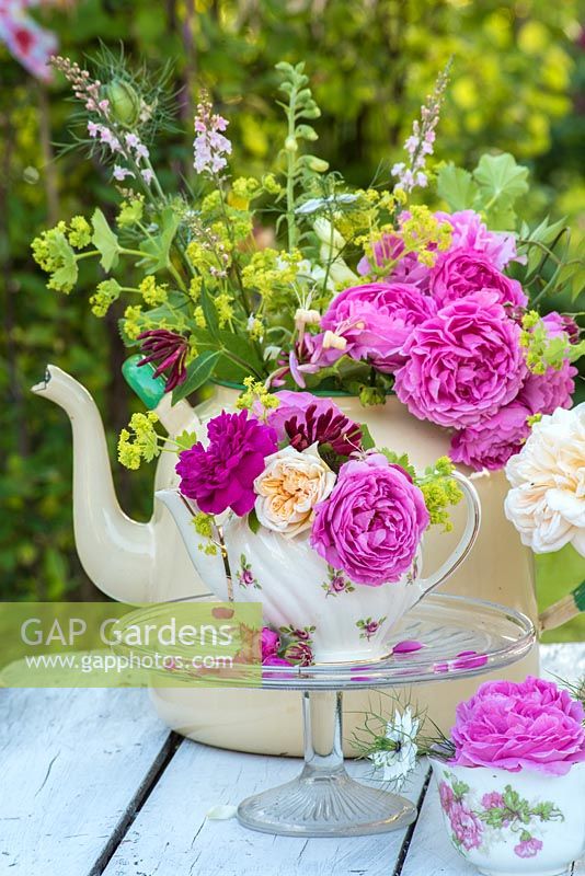 Cut roses arranged in enamel tea set, against a garden backdrop.