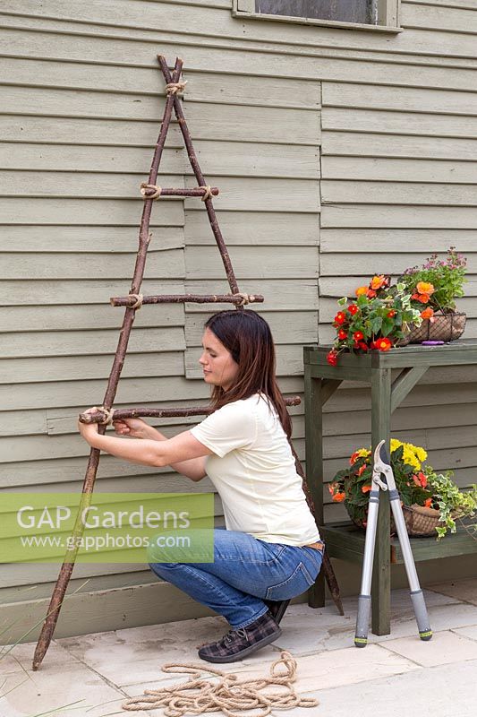 Woman tying horizontal sticks on to the vertical hazel frame to create a ladder for hanging baskets from. 