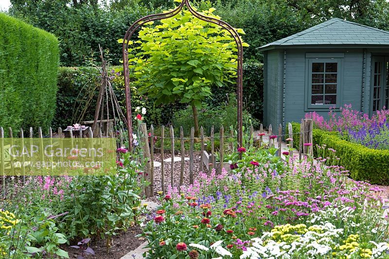View over cut mixed flower beds through metal arch and picket fence to an Catalpa bignonioides - 
Indian bean tree and blue summerhouse