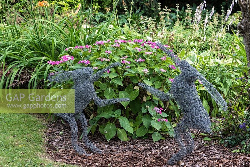 Two dancing hares, crafted from chickenwire, in front of flower border