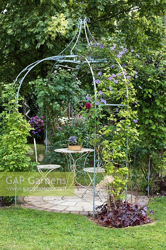 Ornate metal gazebo or arbour above a circular patio with table and chairs. Climbers such as 
 Clematis 'Blue Angel' are trained up outside.
