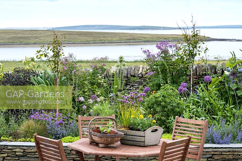 View of wooden table and chairs with view of flowering border and sea. 