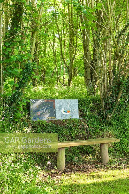 Stone sculptures with engravings behind wooden bench under trees. Plaz Metaxu Garden, Devon, UK. 