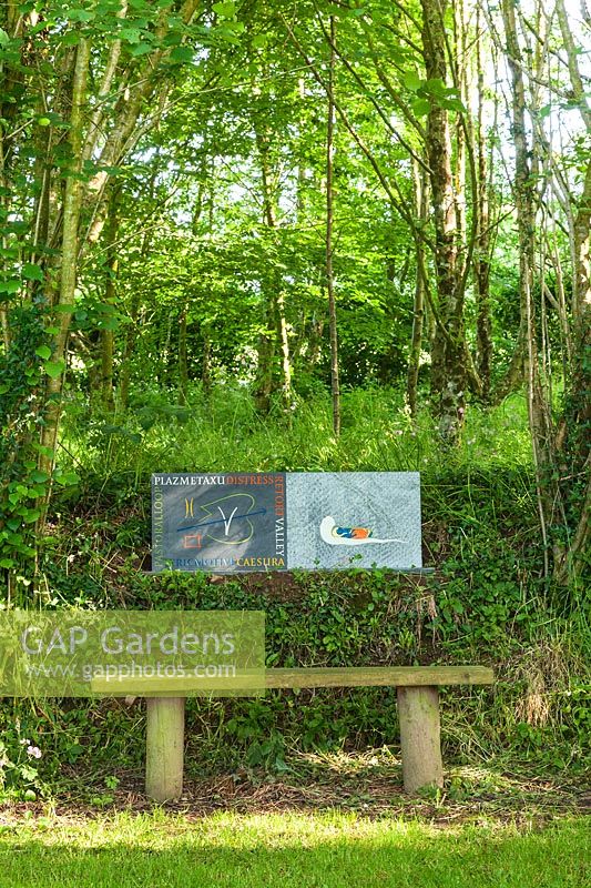 Stone sculptures with engravings behind wooden bench under trees. Plaz Metaxu Garden, Devon, UK.