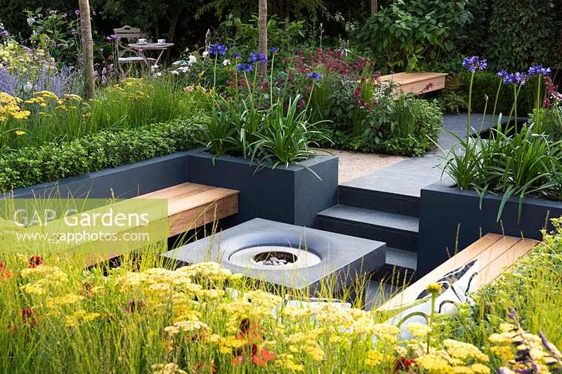 Modern seating area with fireplace surrounded by raised flower beds with 
Achillea, Agapanthus 'Navy Blue' - syn. 'Midnight Star', and Buxus sempervirens.
 Best of Both Worlds garden 
RHS Hampton Court Palace Flower Show 2018