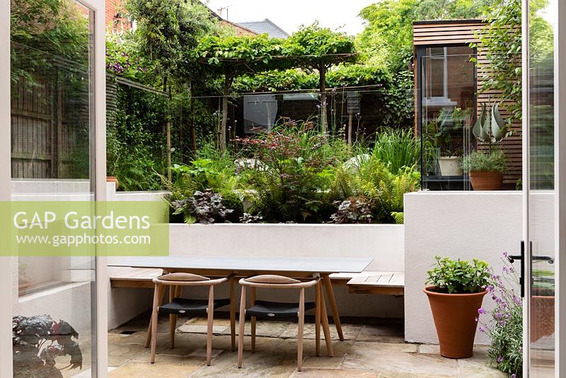 Sunken area in modern, multi-level garden, with cedar panelling, built in seating, white walls, glass railings and steps. 