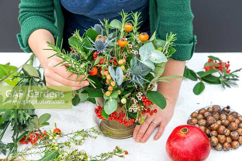 Woman instering Rosemary sprig into mixed festive floral arrangement, with Eucalyptus, Cotoneaster, Hypericum, Erngium, Rose hips and Chamelaucium.