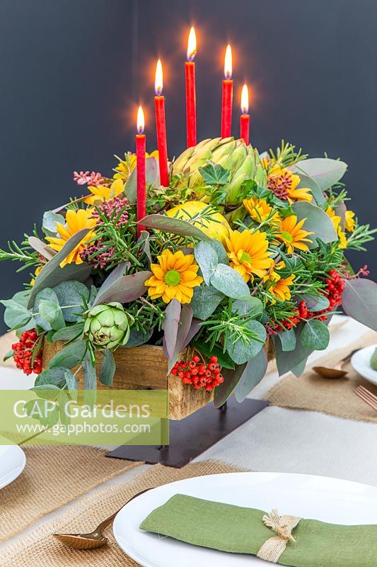 View of completed autumnal floral candle arrangement on set table, with seeded Eucalyptus, Rosemary, Globe Artichokes, Cotoneaster and Chrysanthemum.