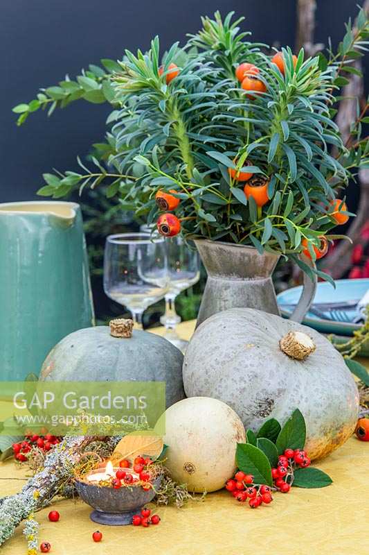 Table centrepiece with galvanised jug of cut Euphorbia foliage and rose hips, harvested squash, moss and candles.