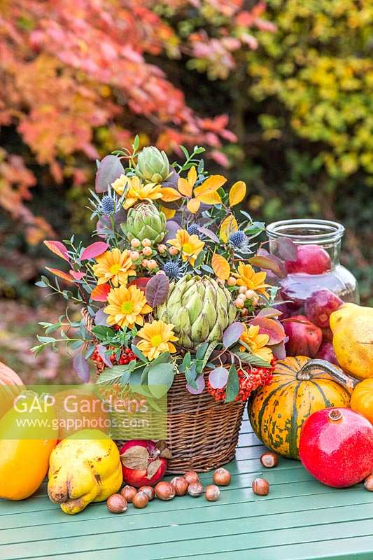 Autumnal floral arrangement in wicker basket, surrounded by harvested vegetables, fruit and nuts.