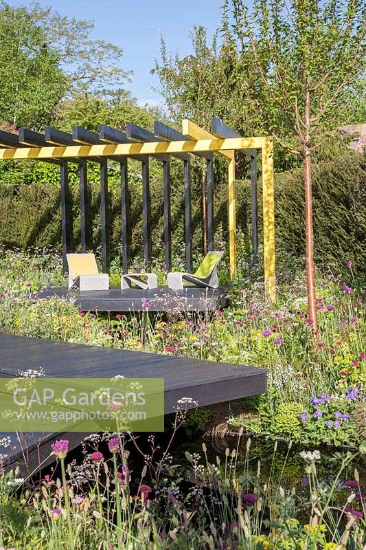 Contemporary, decked seating area under steel-framed pergola, surrounded by mixed naturalistic planting.