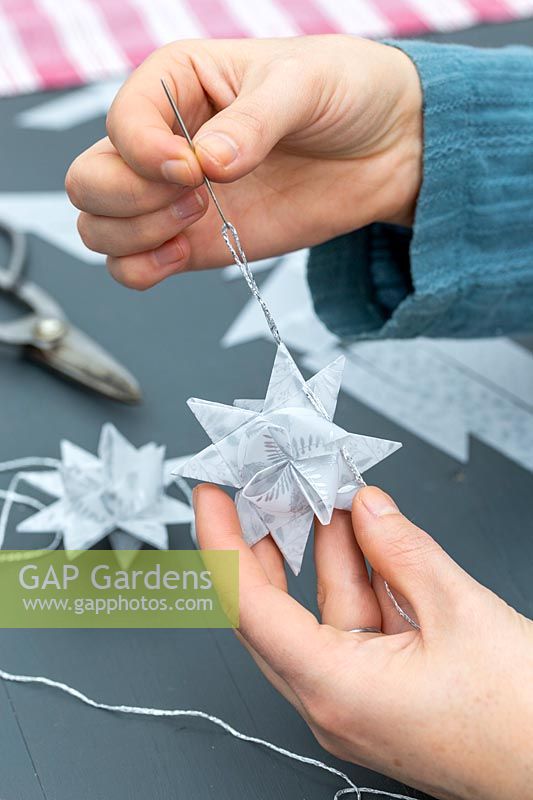Woman using needle and silver thread to create hanging loop on paper star decoration.