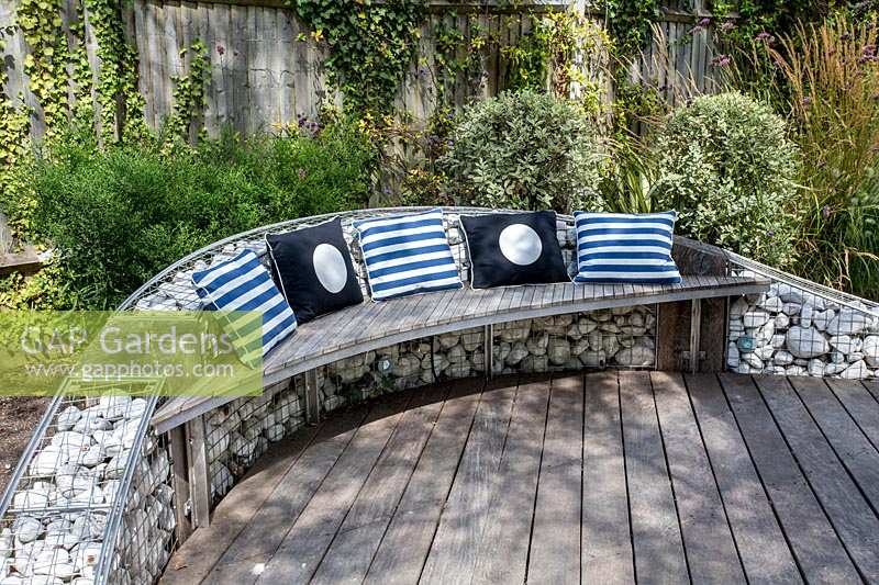 Gabion wall with a hardwood seating attached dressed with garden cushions, 
with wood decking
