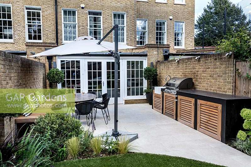 Contemporary back garden with white stone patio, with garden furniture, parasol
 and wooden seating dressed with garden cushions. 
To the right is a garden kitchen and dining area with built-in barbecue.
 In the forground  is an artificial lawn and a bed with Stipa tenuissima
 and Ilex crenata clipped to a ball