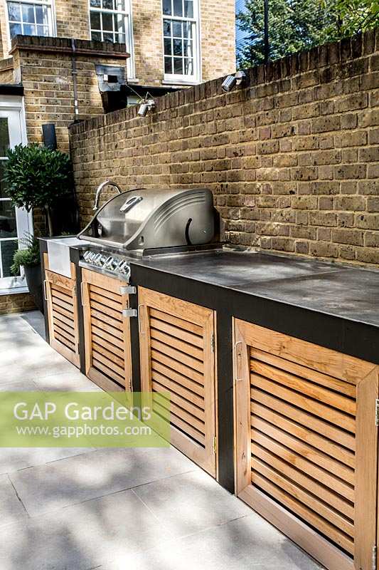 Contemporary garden with white stone patio, with a garden kitchen
 built in barbecue against brick wall.