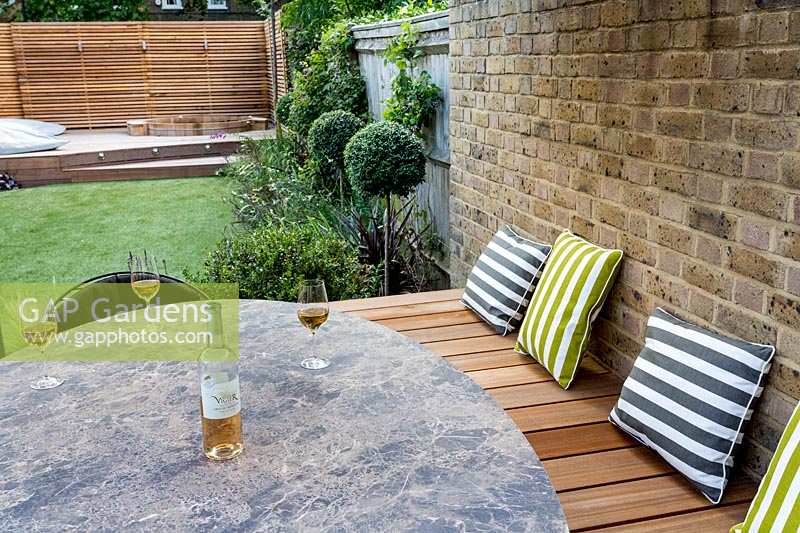 View across modern outdoor table and wooden seating dressed with garden cushions against brick wall
 to an artificial lawn with Ligustrum jonandrum trained as half standards to horizontal wooden fencing