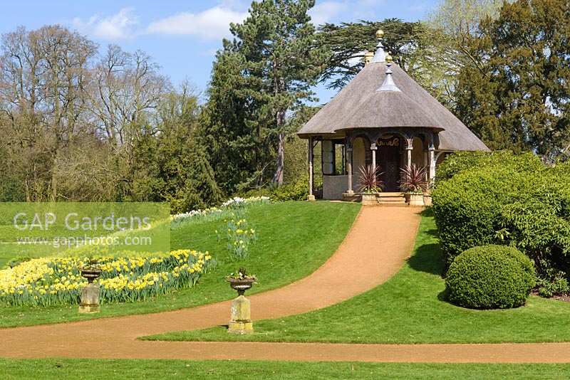 Path leading up to the Swiss-style thatched cottage, areas of lawn planted with drifts of daffodils and shrubs
