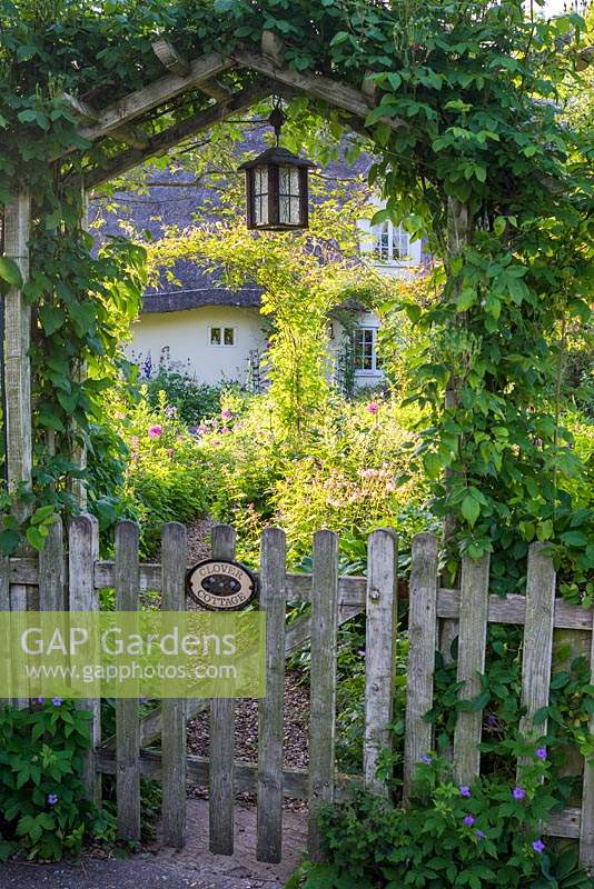 Wooden gateway and fence with view through to cottage garden and cottage. 

