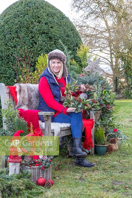 Woman sat on bench holding homemade wreath, surrounded by Christmas greenery and decorations