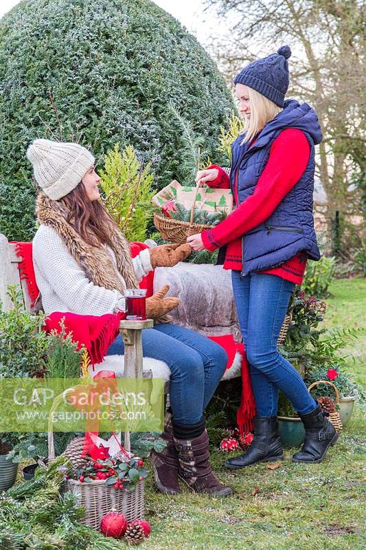 Woman giving basket with presents to friend sat on bench, surrounded by Christmas greenery and decorations