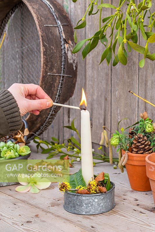 Woman lighting candle in galvanised candle holder decorated with winter flowers, leaves and bark. 