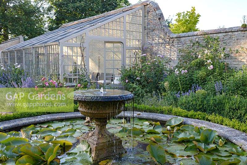 Nymphaea waterlilies in a circular pond with stone fountain surrounded by Buxus - box hedging. Rare Lean-to greenhouse. Rosa 'Queen of Sweden' and Nepeta grandiflora 'Summer Magic' in front of greenhouse