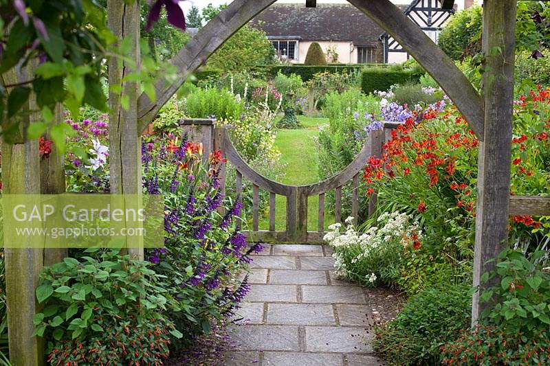 View from under wooden pergola to low gate and flowering herbaceous perennial borders. Wollerton Old Hall, Market Drayton, UK. 
