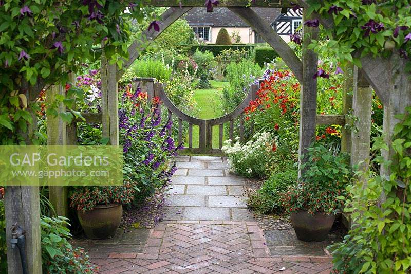 View from under wooden pergola to low gate and flowering herbaceous perennial borders. Wollerton Old Hall, Market Drayton, UK.