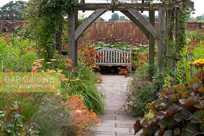 Double, mixed flowerbeds bordering paved pathway, with view to traditional wooden bench. Wollerton Old Hall Garden, Market Drayton, UK.