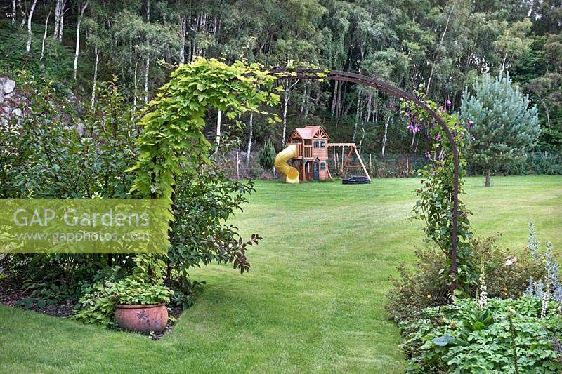 View to children's playhouse under a metal arch covered with climbing Humulus lupulus 'Aureus' and Clematis. 